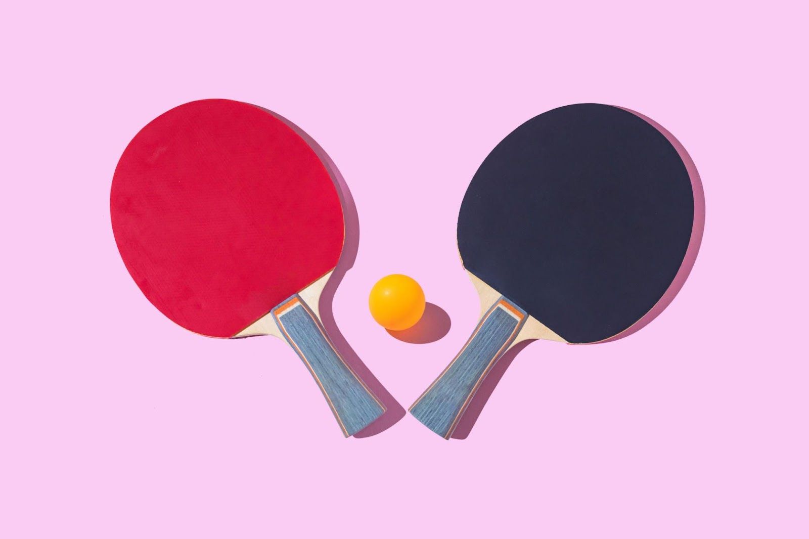 Black and red ping-pong rackets with wooden grip and orange ball on pink background.
